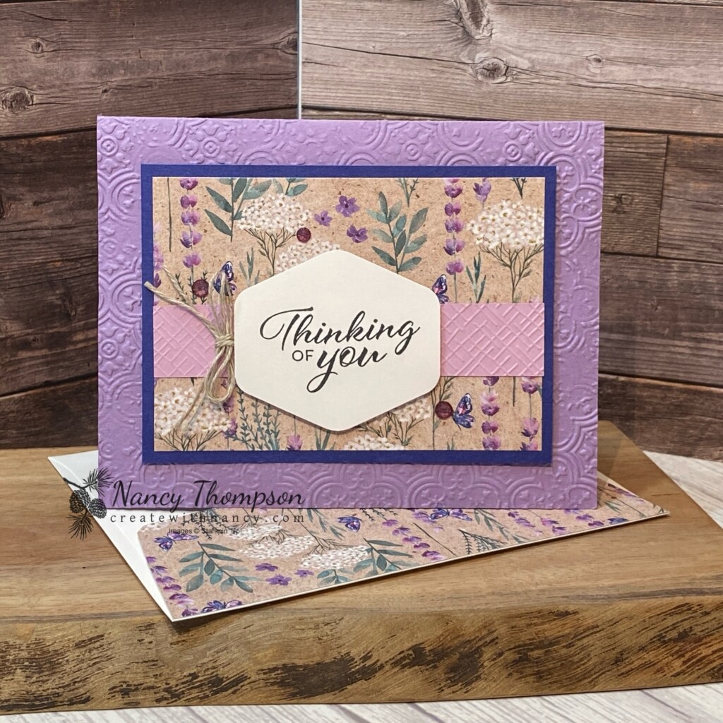 This is a card using the Perennial Lavender designer paper. The card base is a purple color (highland heather) that is embossed with the Distressed Tiles embossing folder. The designer paper in the center of the card is matted onto Starry Sky card stock. There is a strip of Fresh Freesia Card Stock embossed with the Basic 3D (crosshatch) embossing folder in the center with a punched label in the center with the greeting and the words "thinking of you". There is a double tied bow of linen thread on the lift side of the punched label. Purple Fine Shimmer Gems. 