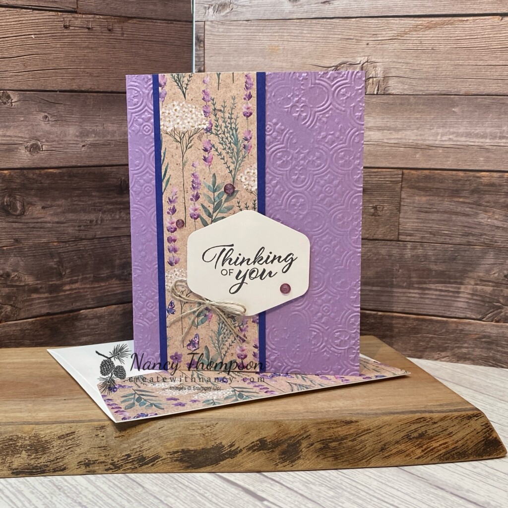 This is a card using the Perennial Lavender designer paper. The card base is a purple color (highland heather) that is embossed with the Distressed Tiles embossing folder. The designer paper in the center of the card is matted onto Starry Sky card stock. There is a strip of Fresh Freesia Card Stock embossed with the Basic 3D (crosshatch) embossing folder in the center with a punched label in the center with the greeting and the words "thinking of you". There is a double tied bow of linen thread on the lift side of the punched label. Purple Fine Shimmer Gems. 