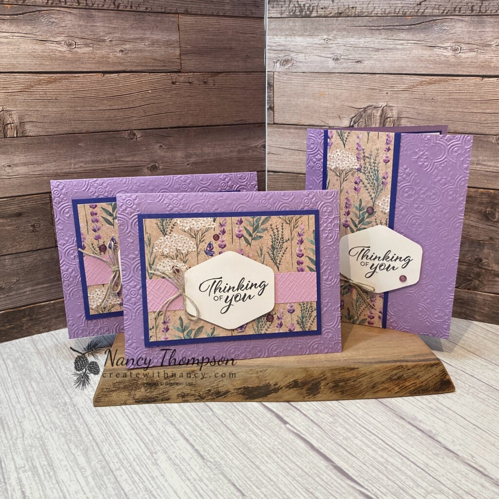 This is a card using the Perennial Lavender designer paper. The card base is a purple color (highland heather) that is embossed with the Distressed Tiles embossing folder. The designer paper in the center of the card is matted onto Starry Sky card stock. There is a strip of Fresh Freesia Card Stock embossed with the Basic 3D (crosshatch) embossing folder in the center with a punched label in the center with the greeting and the words "thinking of you". There is a double tied bow of linen thread on the lift side of the punched label. Purple Fine Shimmer Gems. 